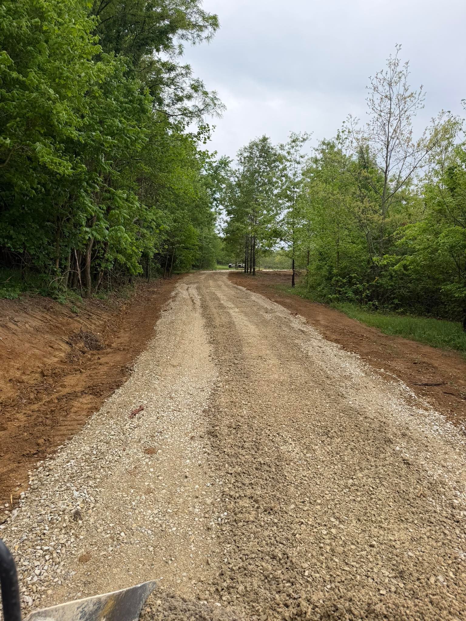 Gravel road through a wooded area under a cloudy sky, with trees lining both sides.