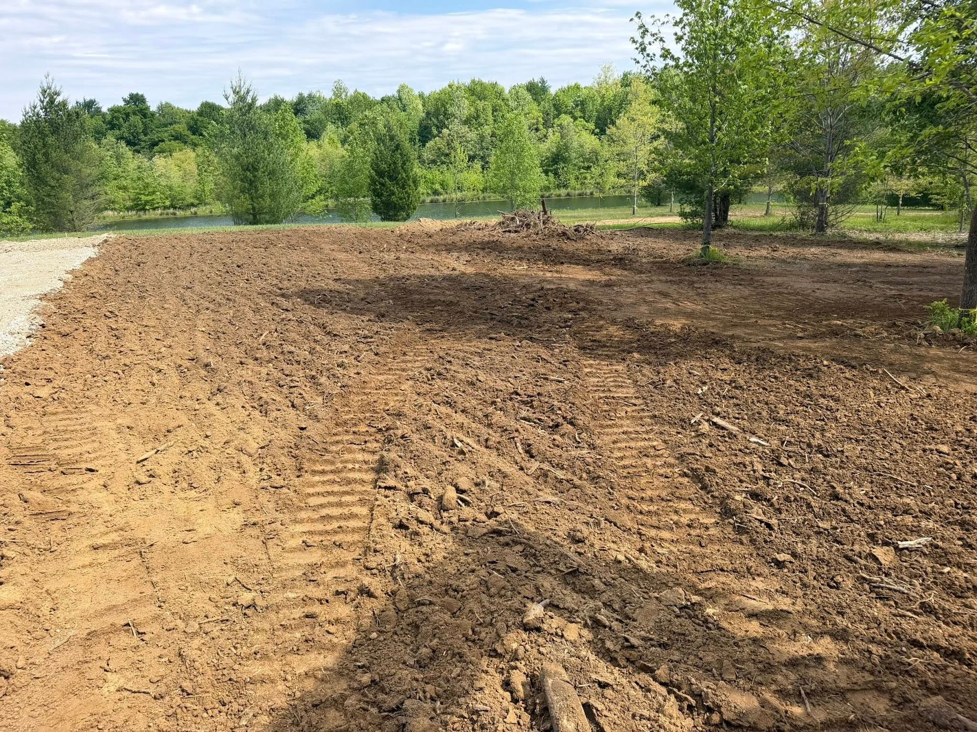 Freshly graded dirt and gravel lot beside green trees under a bright sky
