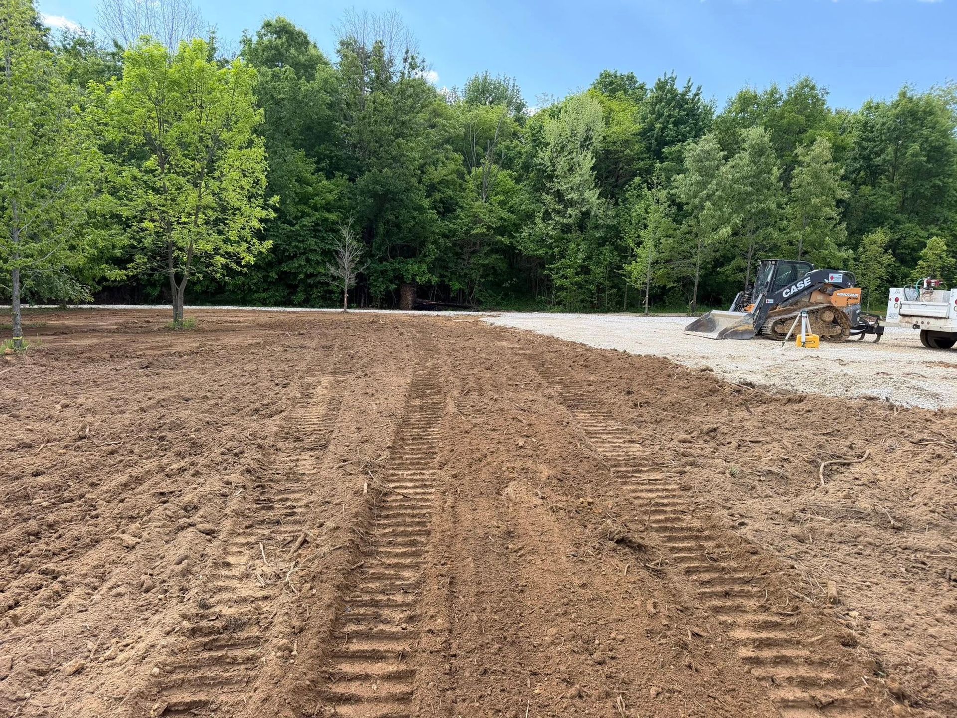 Freshly graded dirt lot with tire tracks, backed by trees and construction equipment