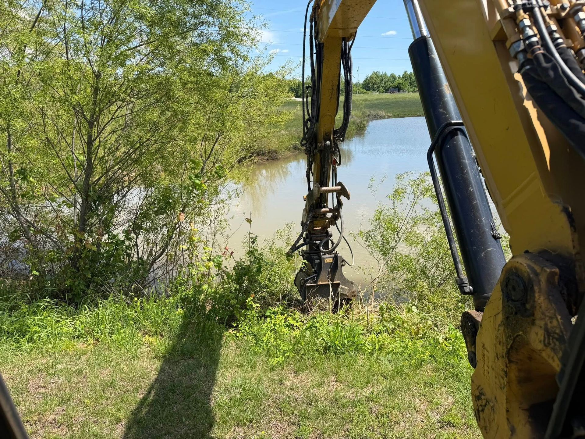 Yellow excavator arm clearing brush by a lakeside with trees and tall grass nearby