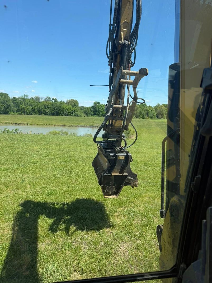 Excavator arm with grappler hovering over a green field beside a pond and trees