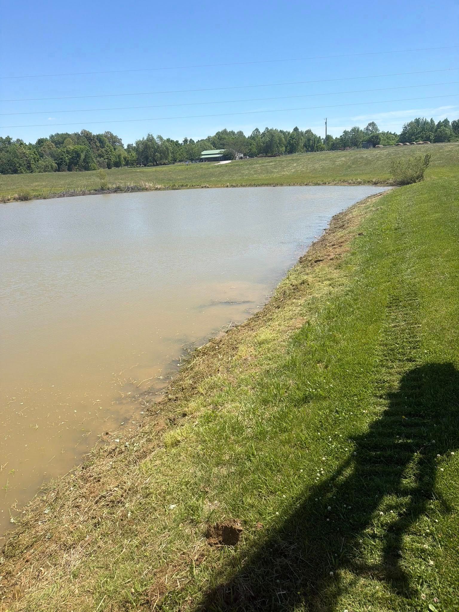 Curved muddy riverbank with grassy shore under a clear blue sky, with a shadow in the foreground
