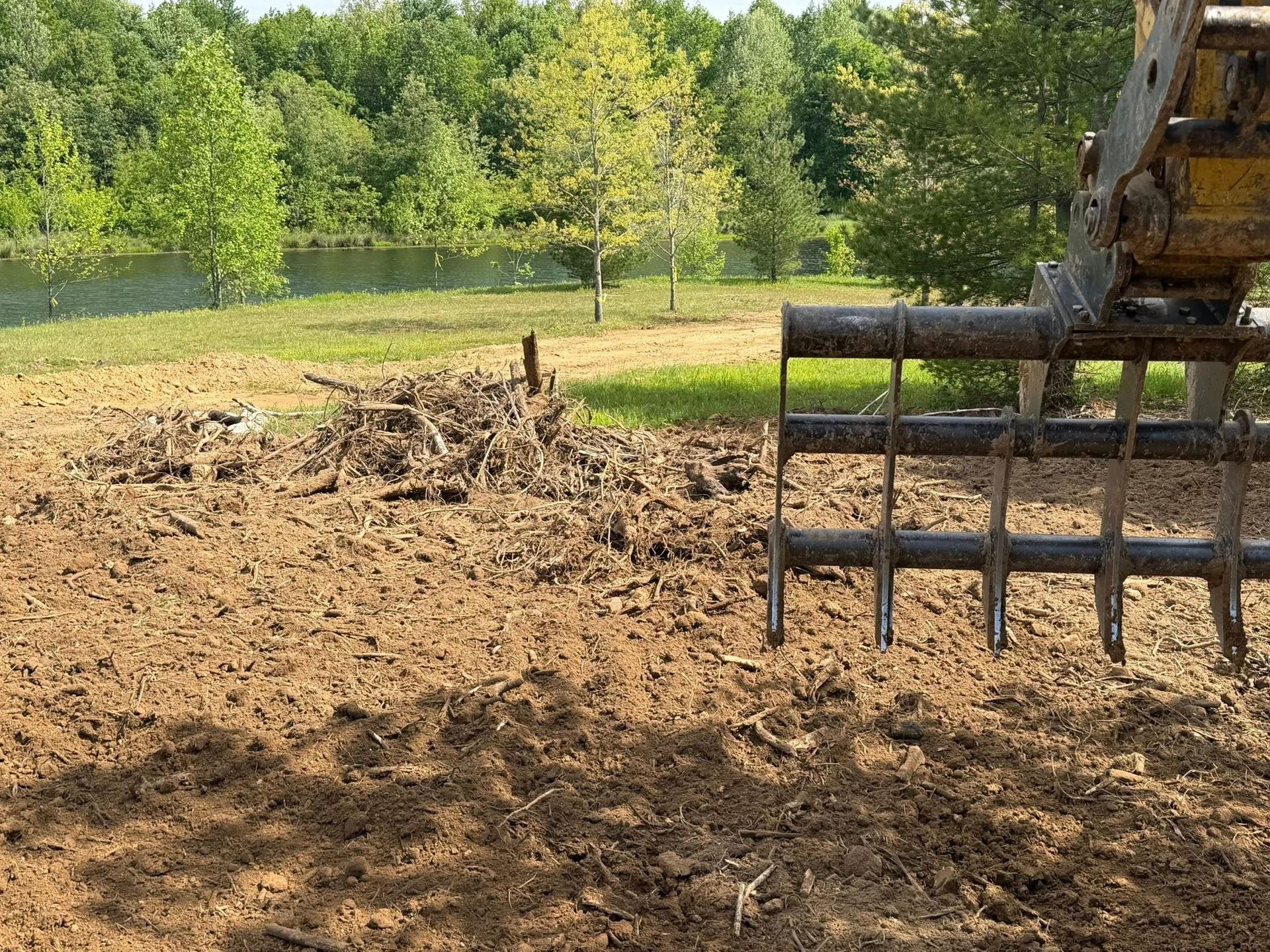 Excavator bucket beside a pile of dirt at a muddy work site with trees and a pond in the background
