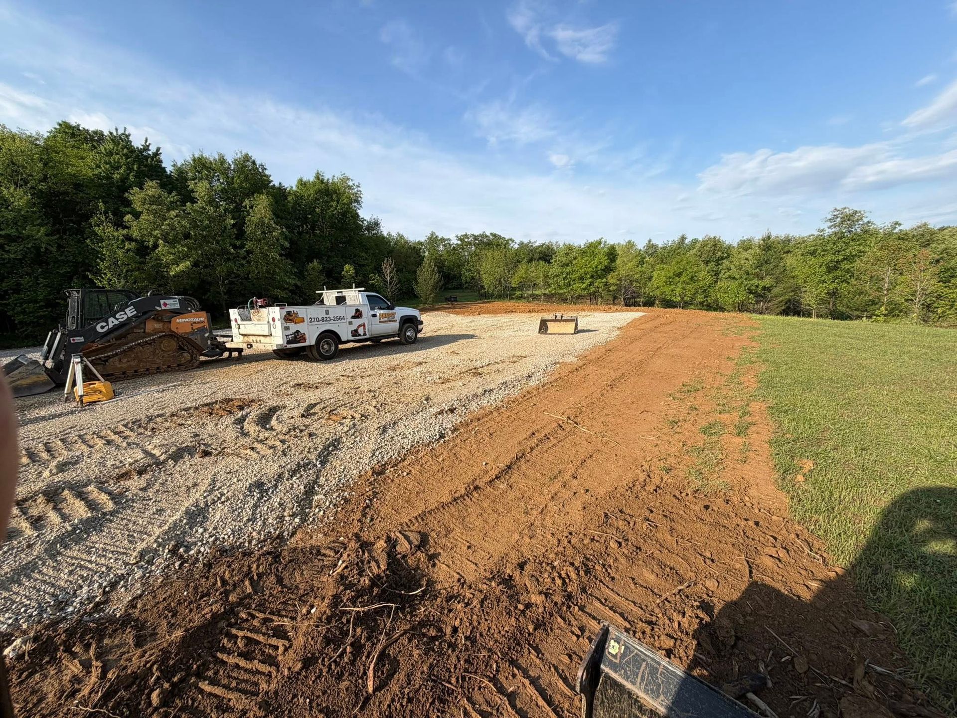 Construction site with gravel road, white truck, dirt piles, and trees under a clear blue sky