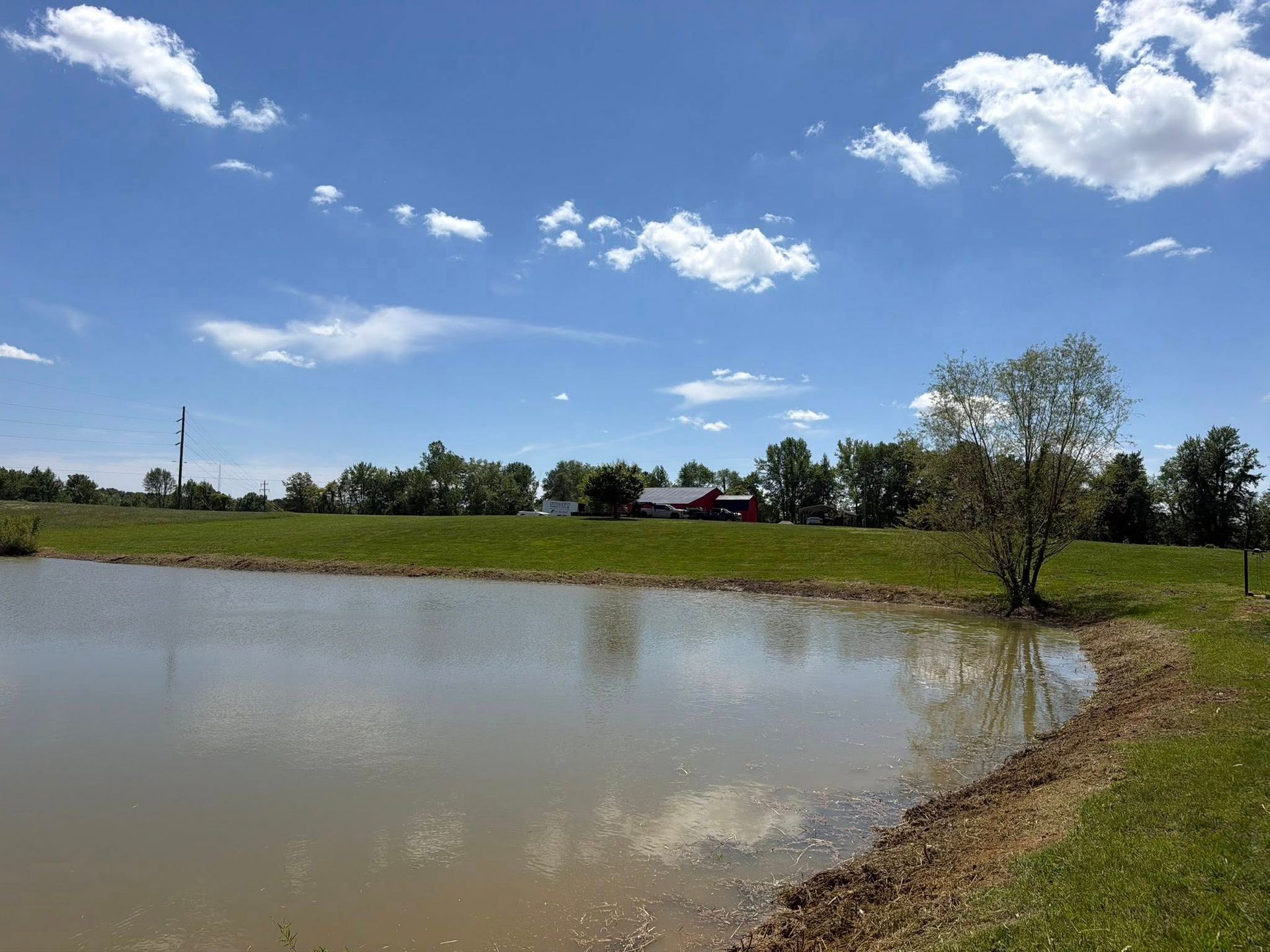 Flooded field under a bright blue sky with clouds, trees, and a dirt shoreline.