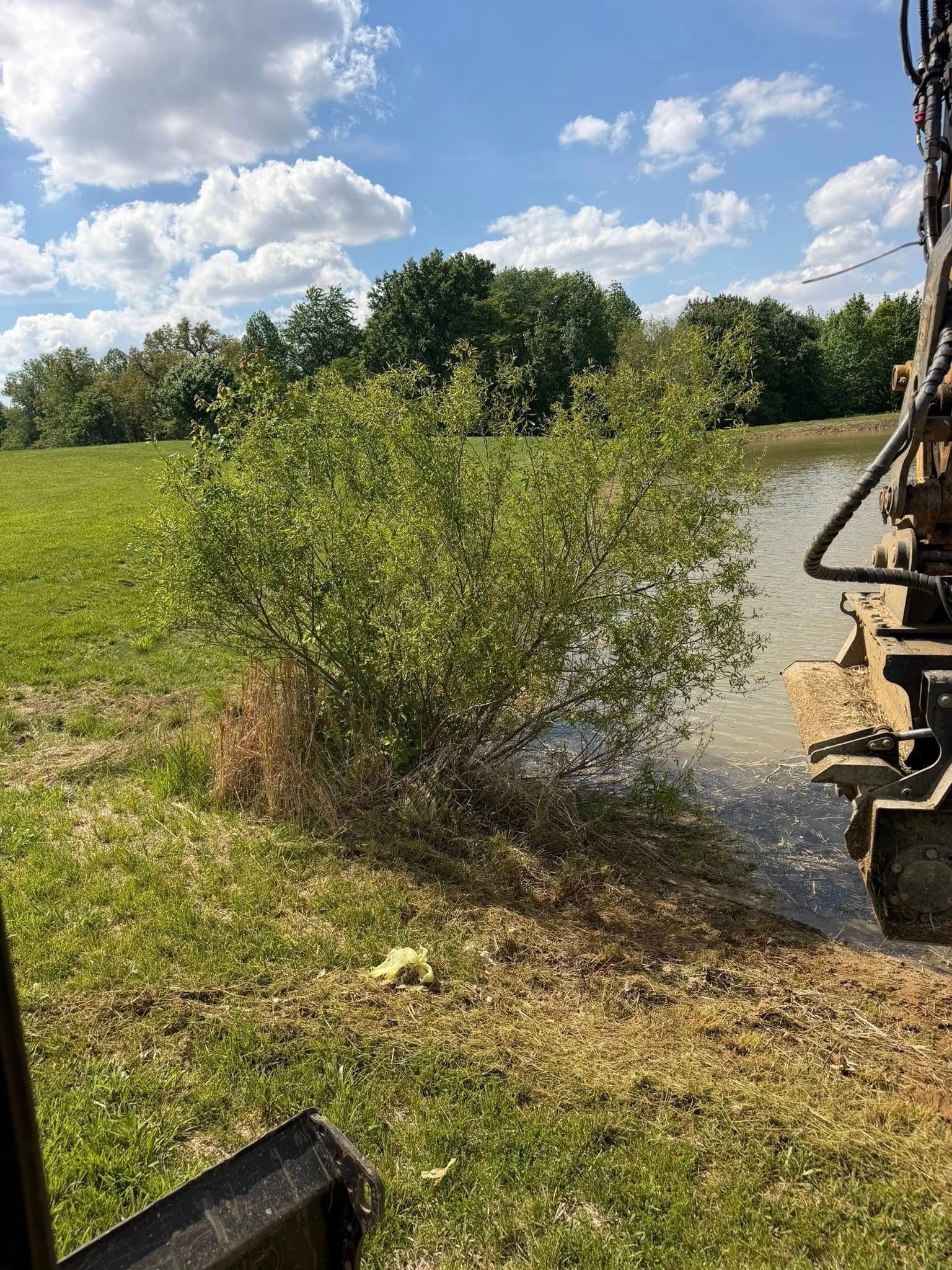 Bulldozer spraying muddy water at bushes beside a grassy field under a blue sky
