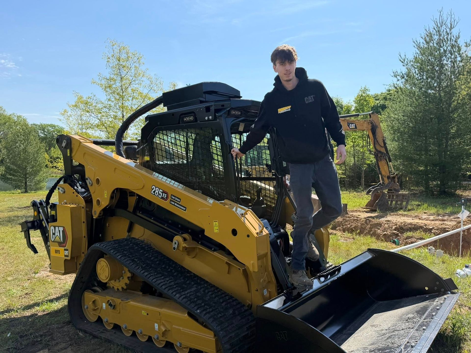 Person standing on a yellow compact skid steer outdoors on a sunny day