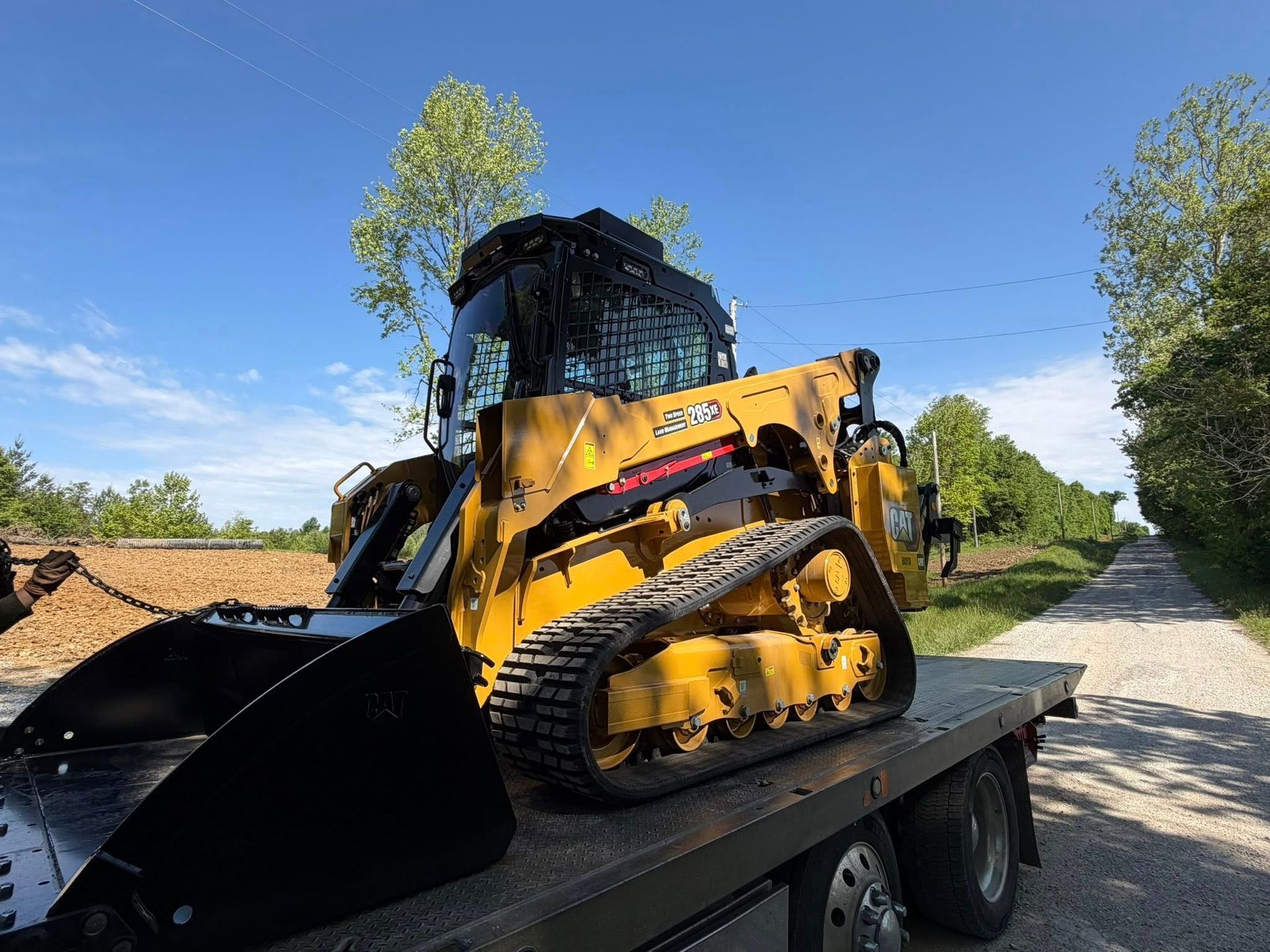 Yellow compact track loader on a flatbed trailer beside a gravel road under a blue sky.