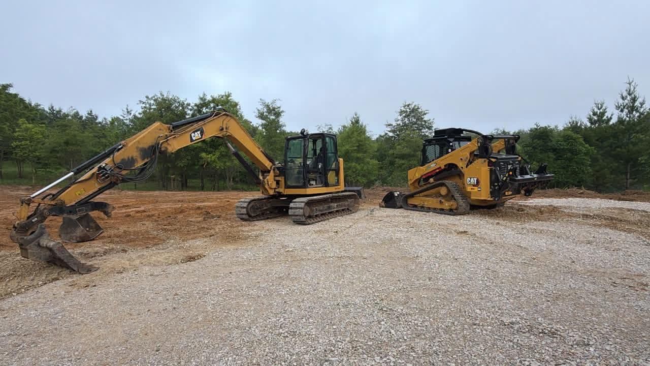 Two excavators on a gravel lot, one digging and one parked, with trees in the background