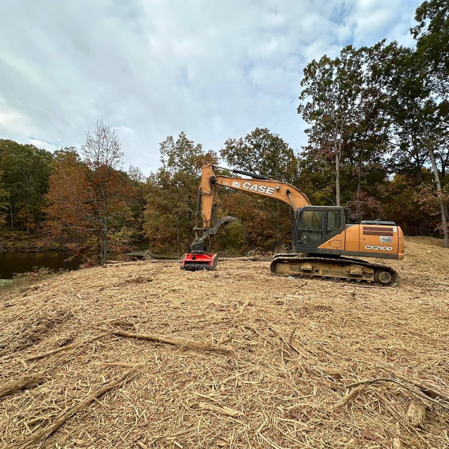 Orange excavator clearing wood chips in a wooded area under an overcast sky.