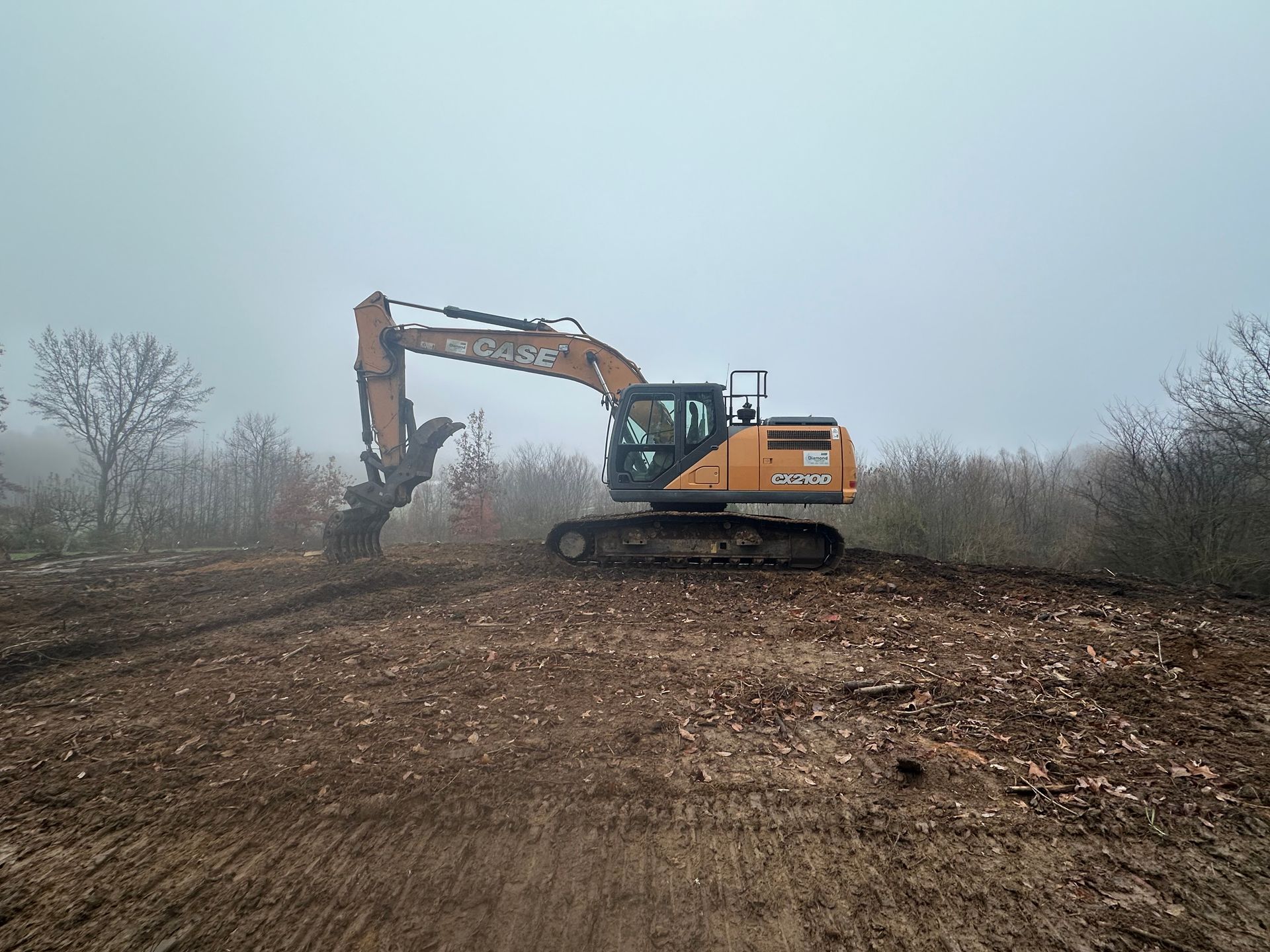 Yellow excavator clearing a foggy dirt lot beside leafless brush