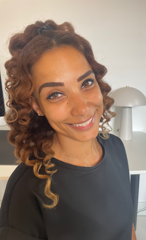 Foto vanFinch Logopediepraktijk, Een vrouw met krullend bruin haar lacht naar de camera. Ze draagt ​​een zwarte blouse en staat in een kamer met een lamp en een wit bureau.