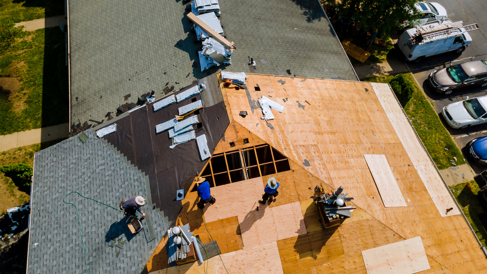 Aerial view of workers replacing shingles on a house roof, showing sections of exposed plywood decking and new shingles.
