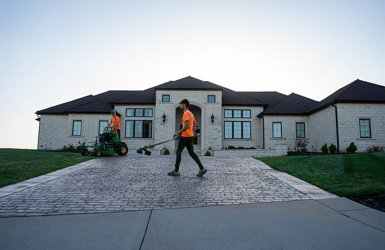 Two people mowing the lawn of a large, light-colored house.