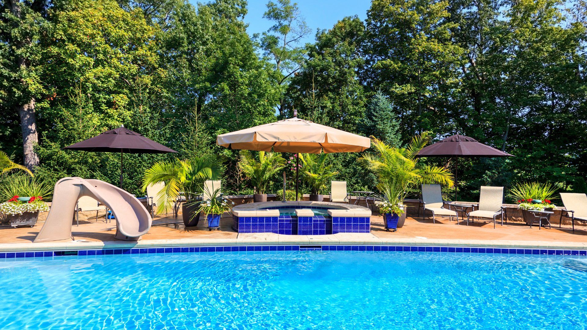 Beautiful backyard pool with blue tiling. There is a slide, jacuzzi, umbrellas and patio furniture. The pool deck is a made from concrete.