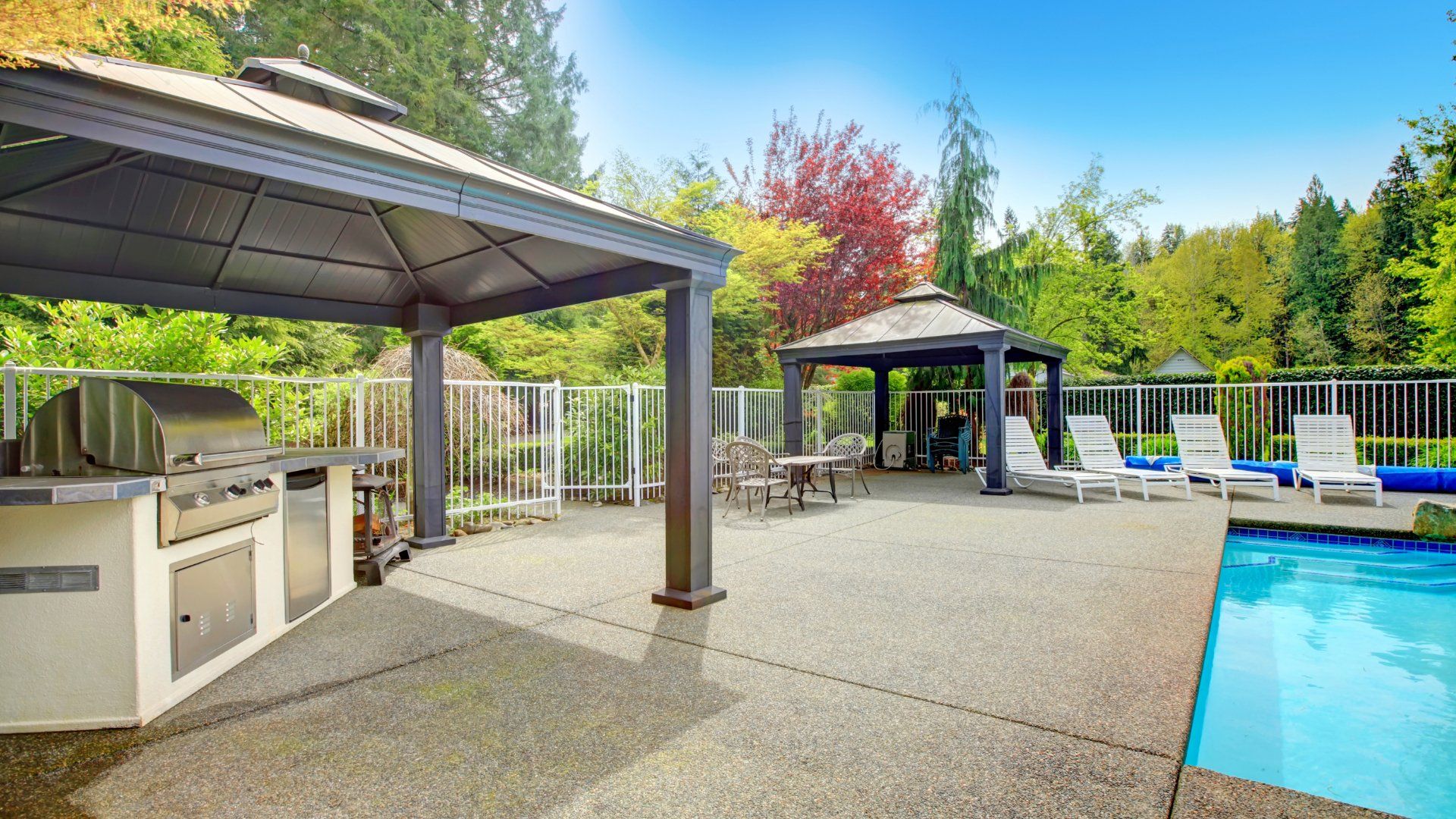 Backyard pool with some sheltered areas. The backyard features a white metal fence, a barbeque area, patio furniture and a stamped concrete pool deck.