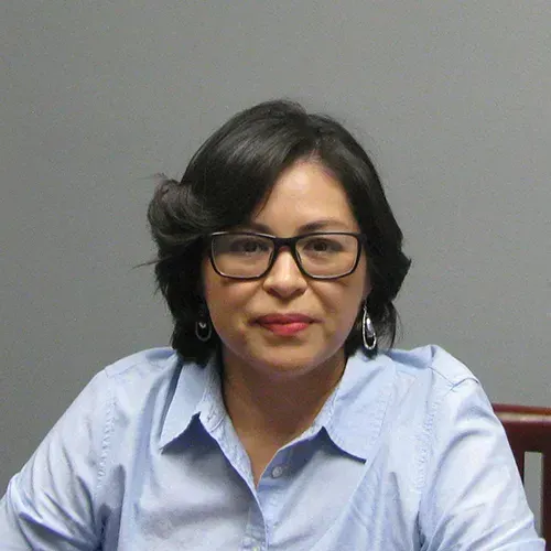 Woman wearing glasses and a light blue shirt, seated against a gray wall, looking at the camera.
