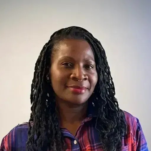 Portrait of a smiling person with long braided hair wearing a purple plaid shirt against a plain wall