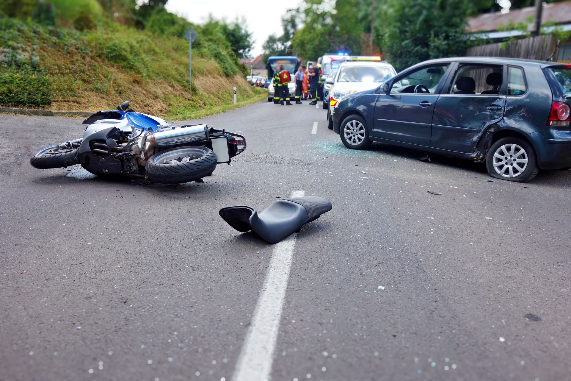 Motorcycle and black car crashed on a road with debris and emergency vehicles in the background