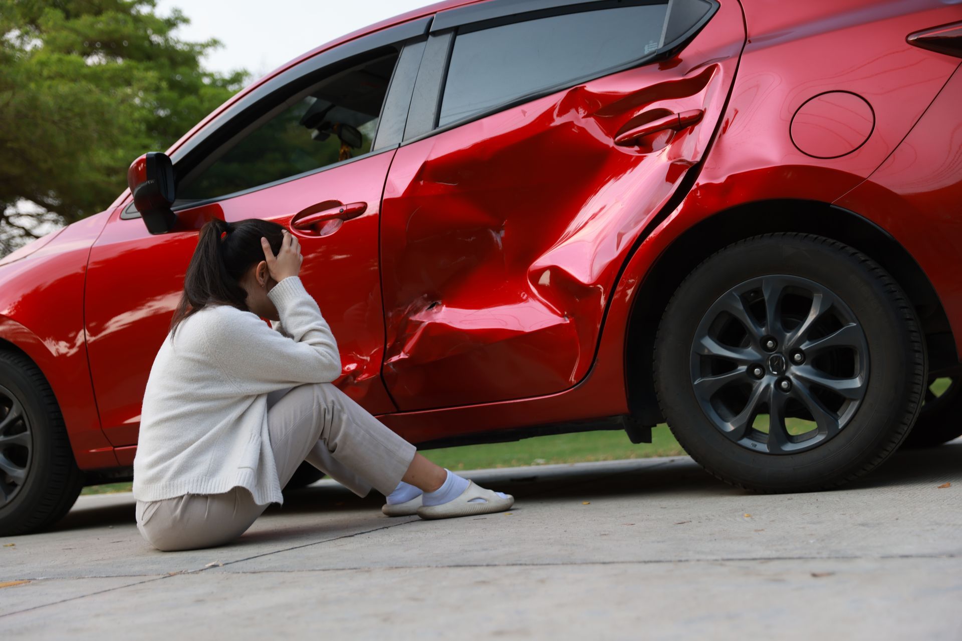 Person crouches beside a damaged red car, inspecting a large dent in the side.