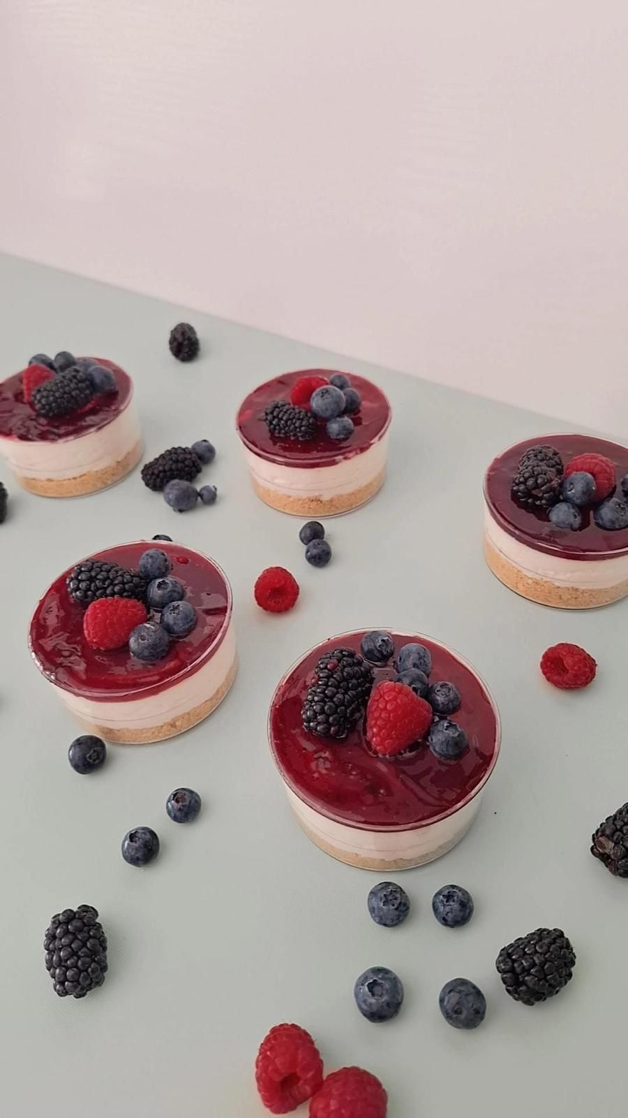Mini cheesecake con frutti di bosco su una superficie grigio chiaro, guarnite con lamponi e mirtilli sparsi.