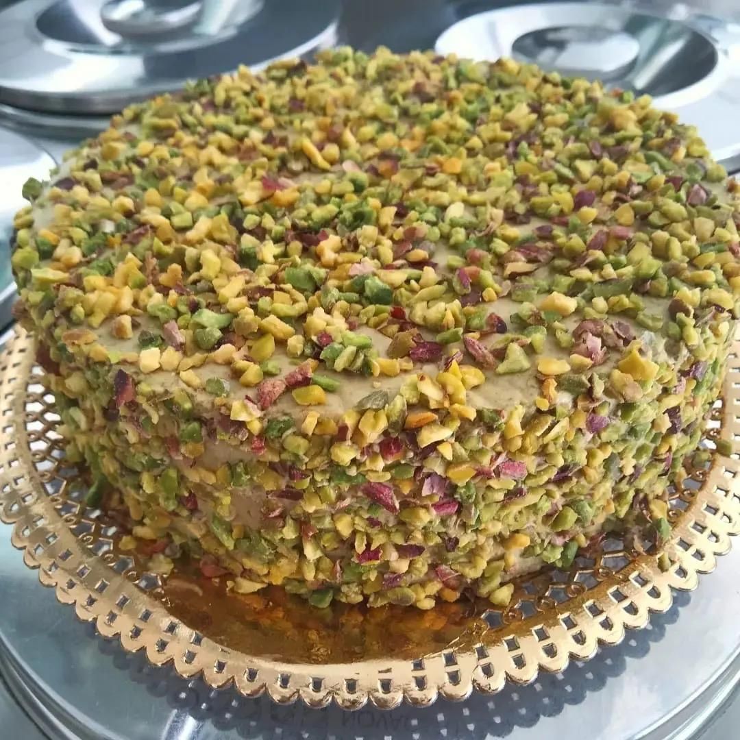 Torta rotonda ricoperta di pistacchi, adagiata su un vassoio decorativo, con la parte superiore e i lati cosparsi di pistacchi tritati.