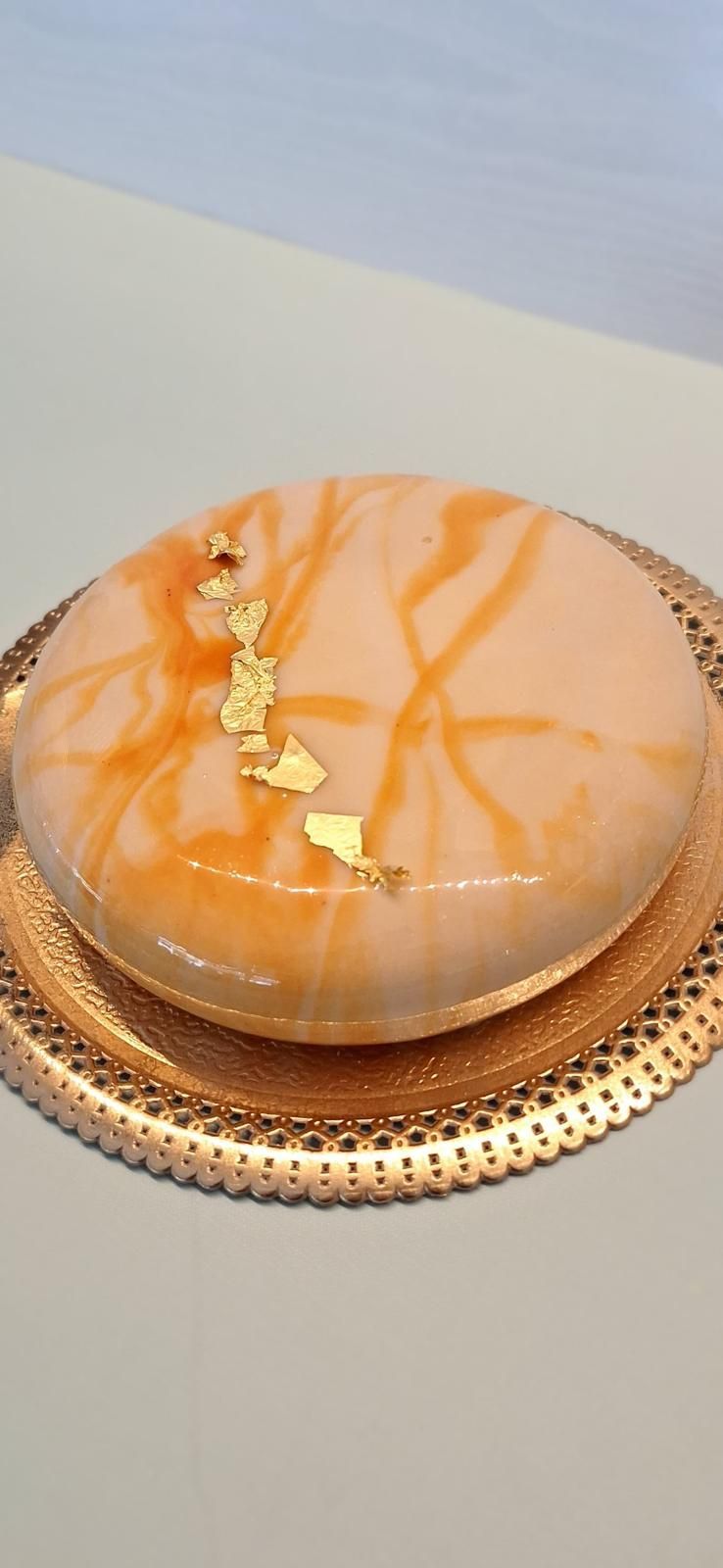 Dessert rotondo glassato alla pesca, servito su un piatto con bordo dorato e una decorazione a filo d'oro.