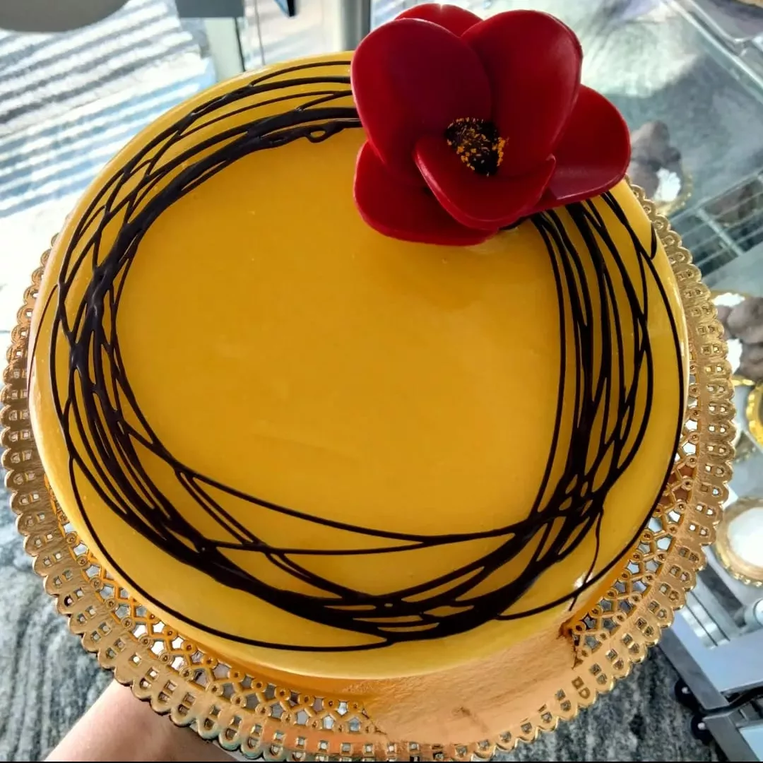 Torta rotonda gialla con glassa al cioccolato e decorazione a fiore rosso, servita su un vassoio decorativo.