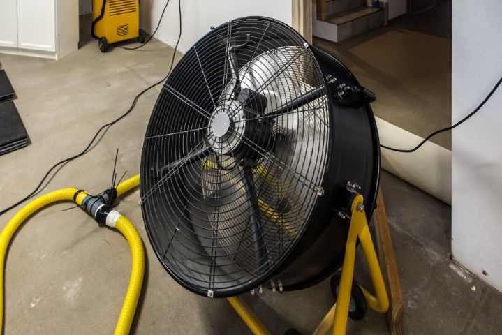 Large industrial fan with black blades and yellow stand, connected to a yellow hose in a room.