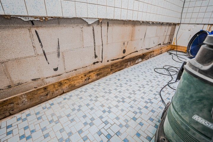 Interior demolition; exposed cinder block wall, damaged trim, blue-white tiled floor, tools, and a fan.