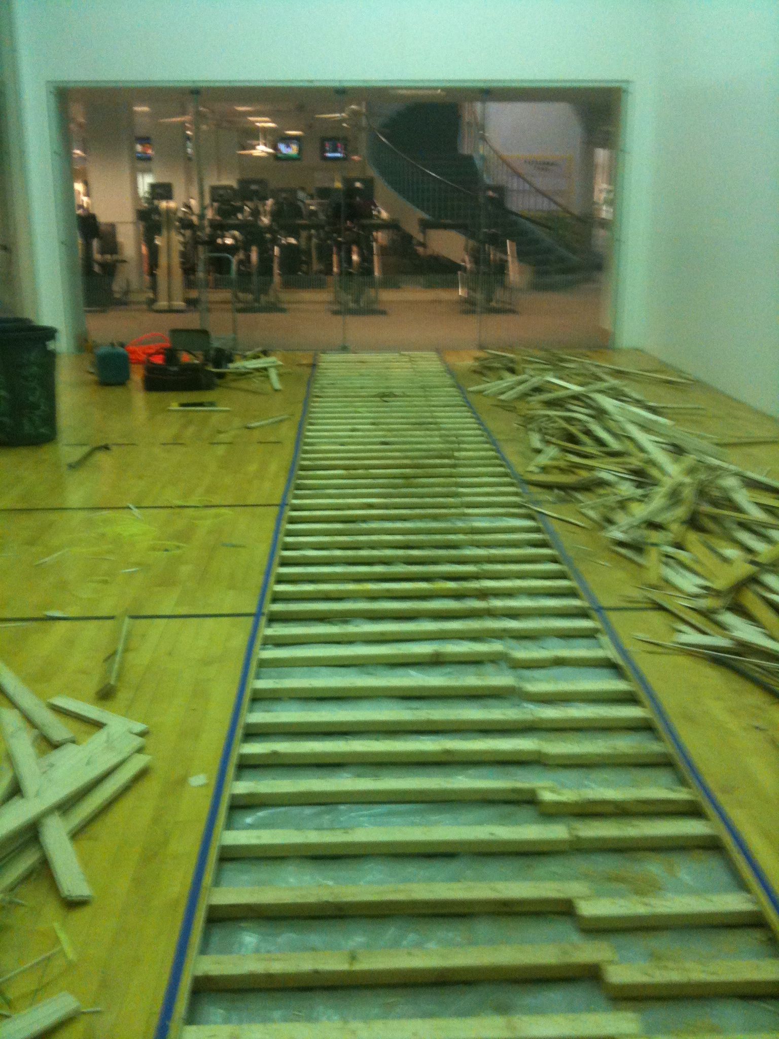 Long metal ladder laid across a gym floor toward a mirrored wall, with exercise equipment in the background