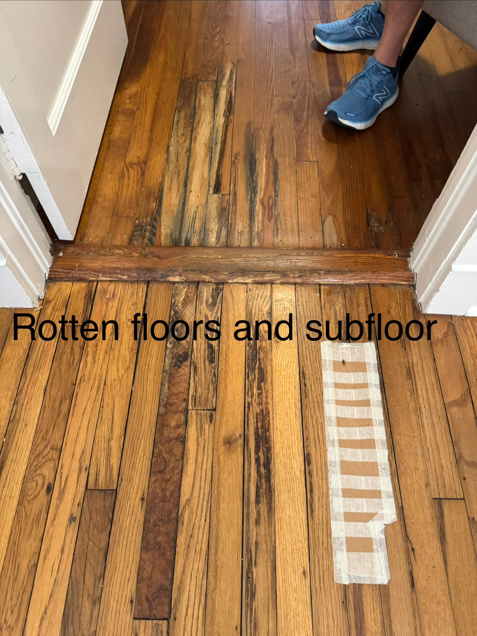 Damaged wooden floor and subfloor in hallway, with a person’s blue shoes visible.