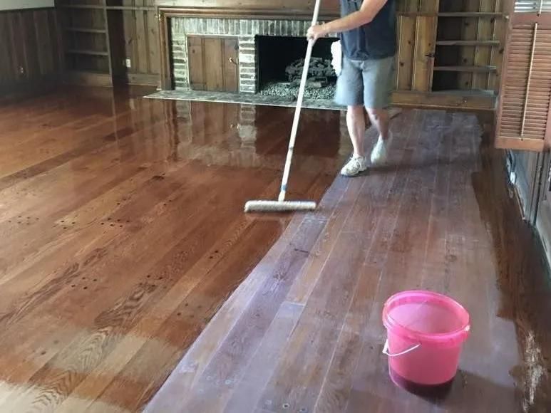 Person mopping a shiny wooden floor with a pink bucket nearby in a room with a fireplace