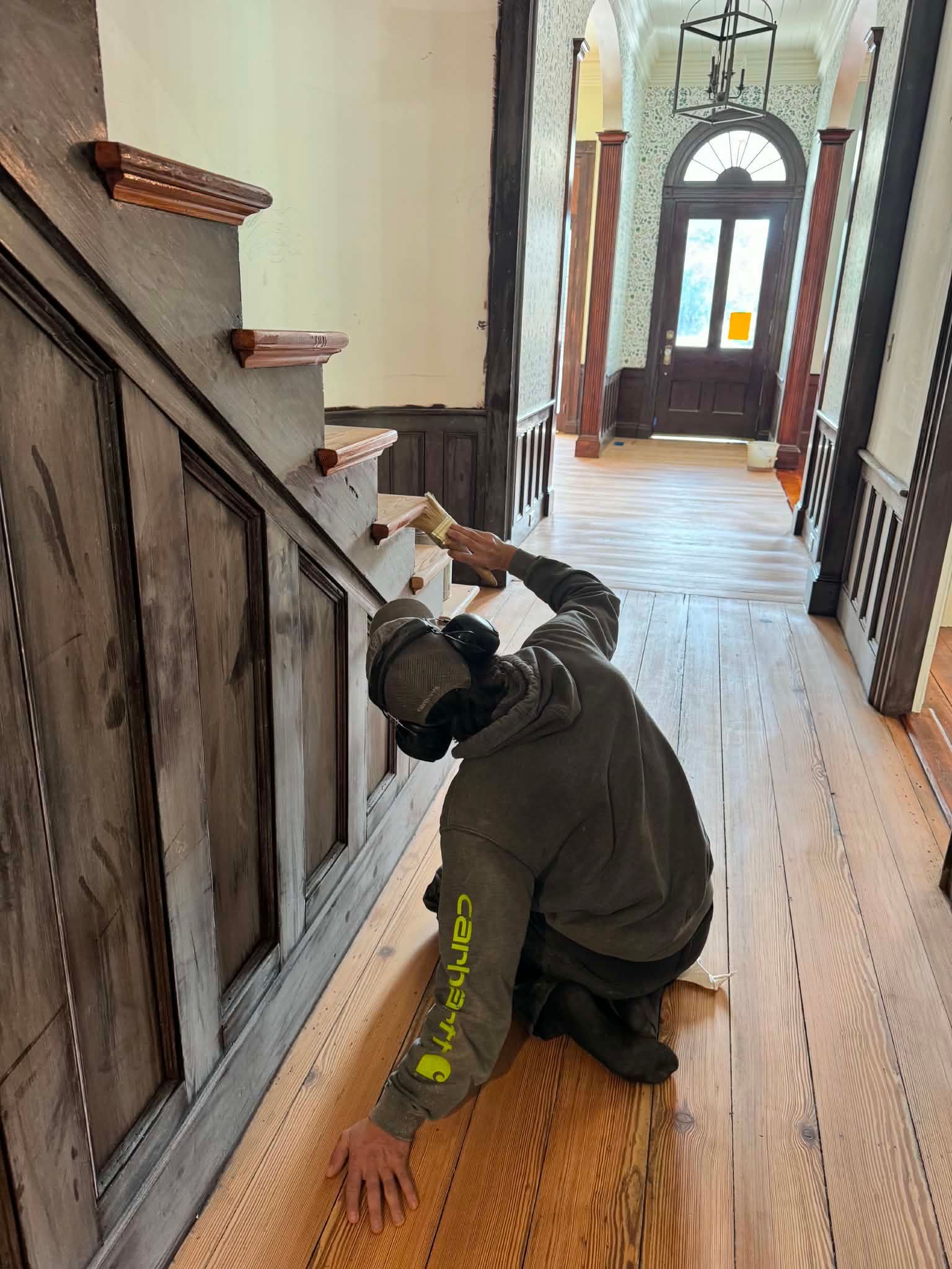 Person kneels on a wooden floor in a hallway, installing trim beside a staircase.