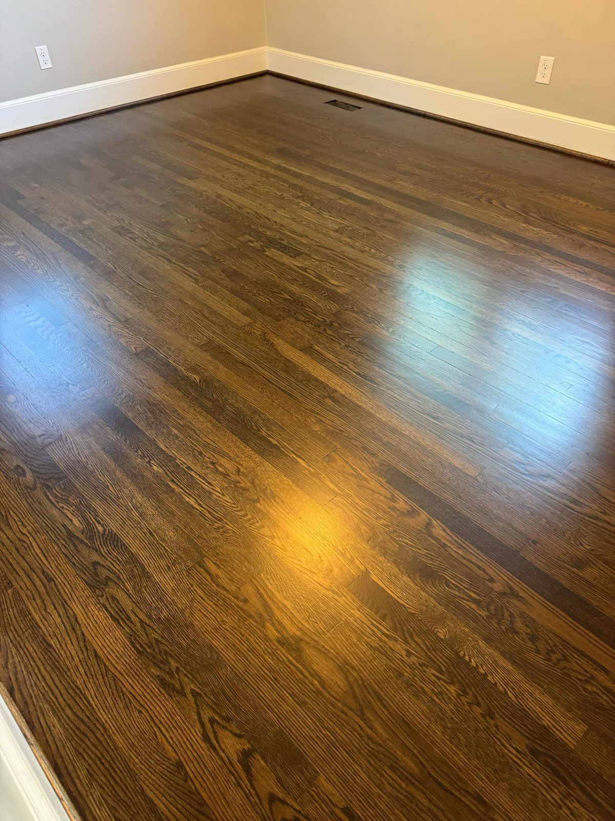 Empty room with dark wood flooring, beige walls, and sunlight reflections on the floor.