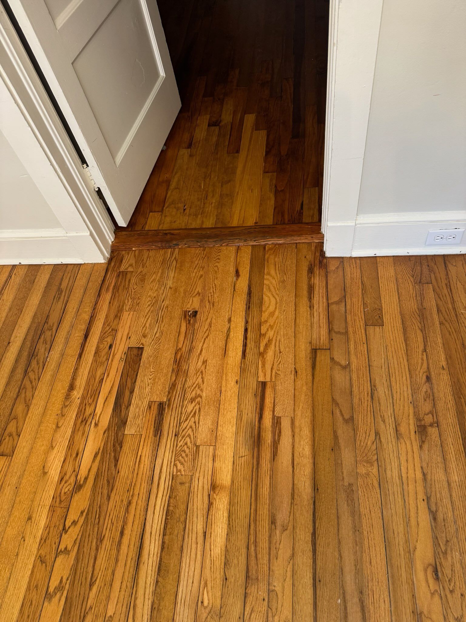 Wooden hallway floor with a damaged seam and dark stain near a doorway, beside white baseboards.