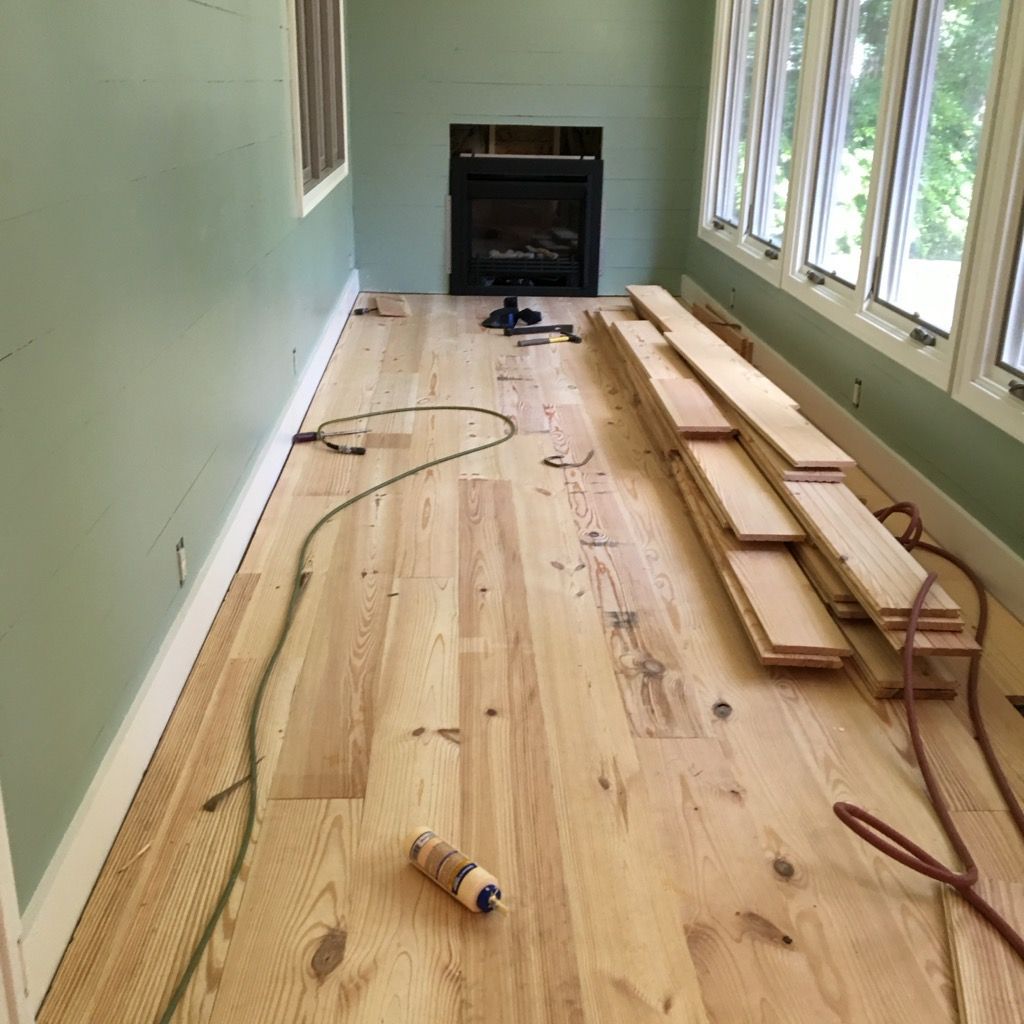 Long narrow room with unfinished wood floorboards stacked along one wall and a fireplace at the end.