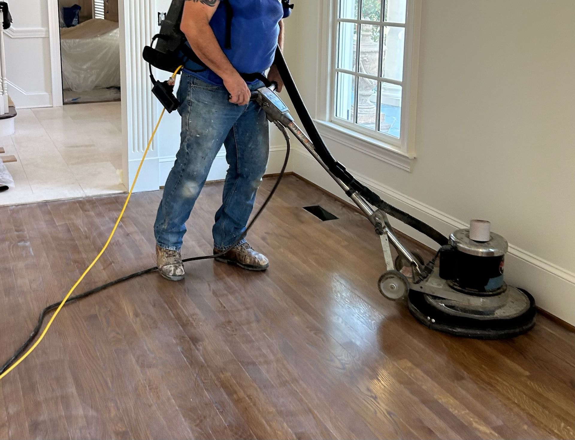 Worker sanding a wooden floor with a floor sander near a window.