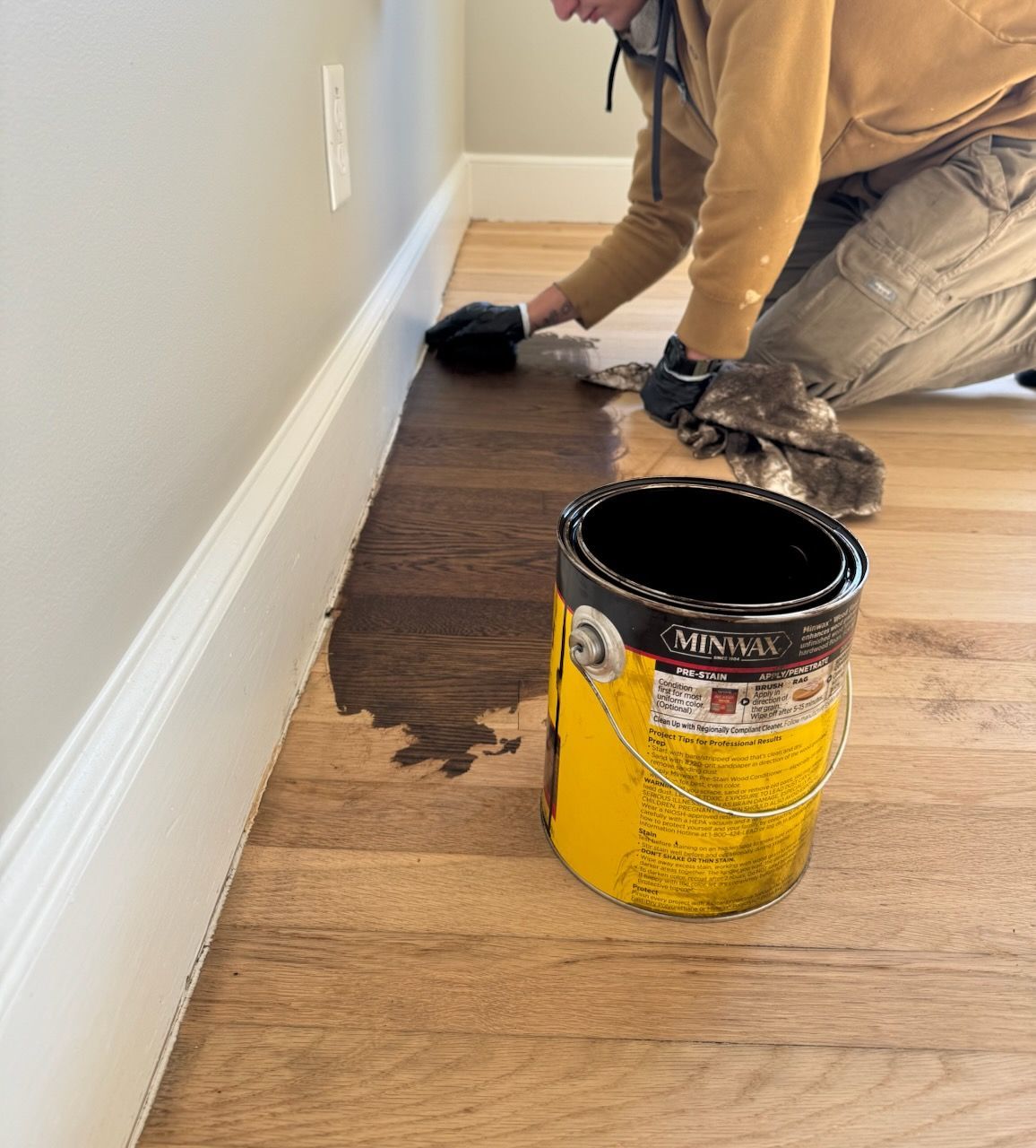 Person painting a floor edge beside a wall with a yellow paint can in the foreground