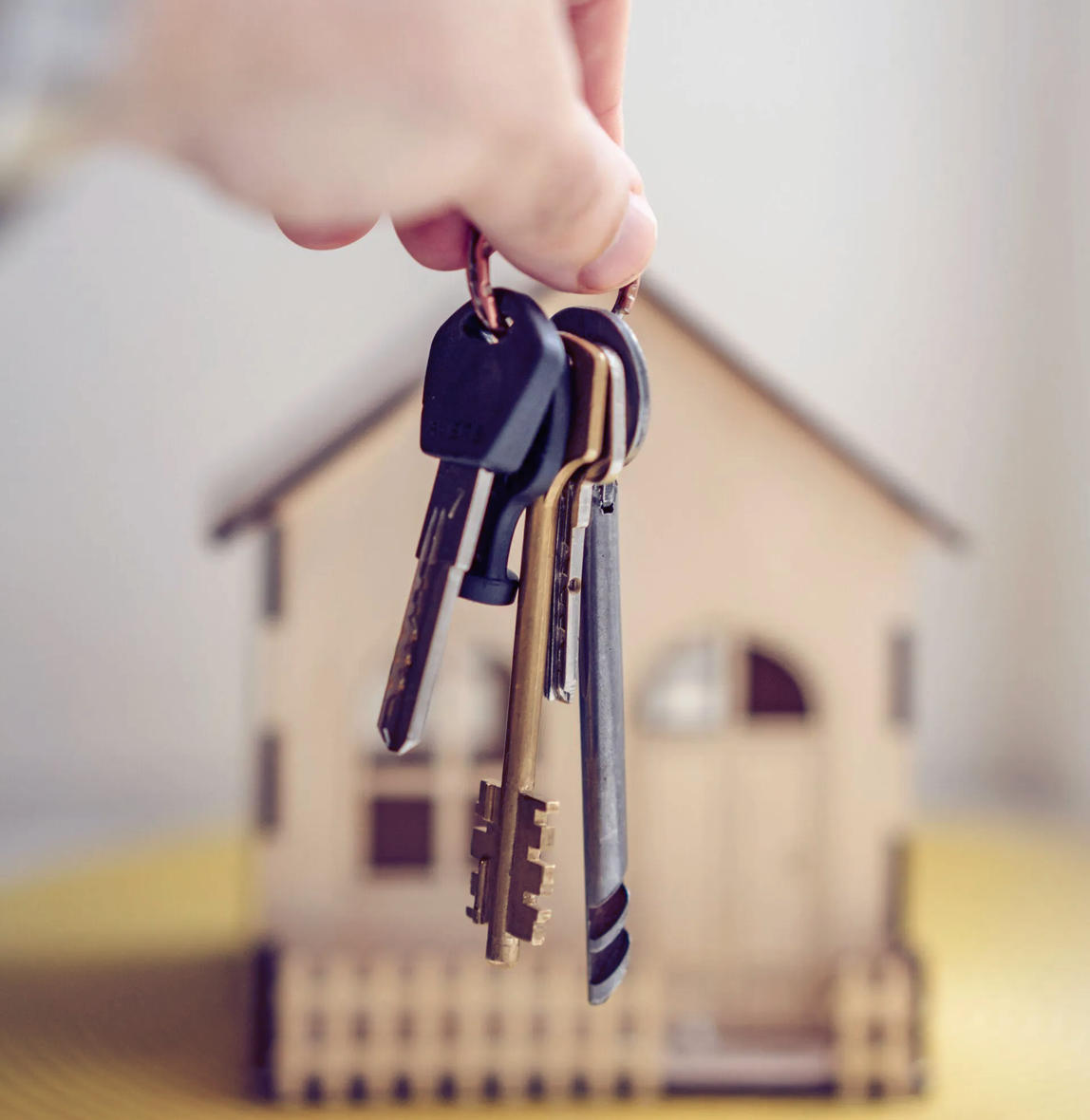 Hand holding house keys in front of a small wooden house cutout.