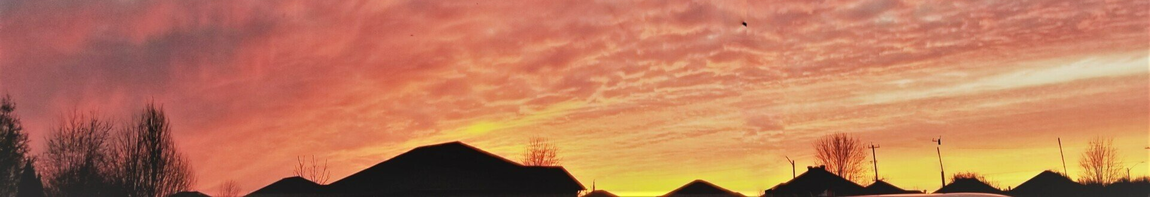 Sunset with fiery red, orange, and yellow sky over a dark silhouette of trees and buildings.