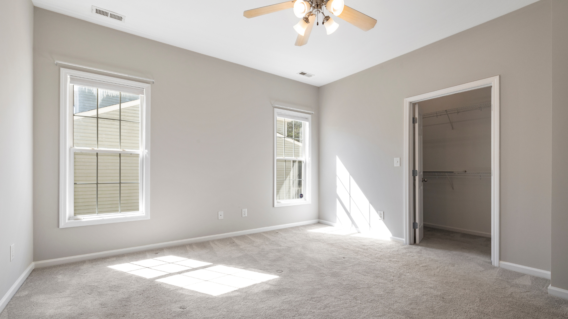 Empty bedroom with beige carpet, light gray walls, two windows, and an open closet.