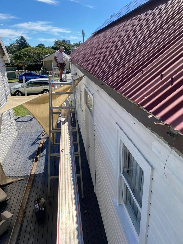 A Man Is Standing On A Ladder On The Side Of A Building — The Guttering Cleaning Man in Forster, NSW