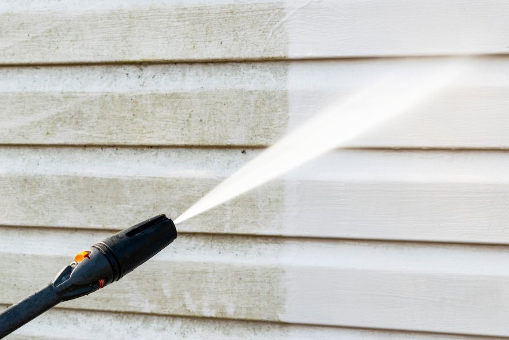 A Person Is Cleaning The Side Of A House With A High Pressure Washer — The Guttering Cleaning Man in Old Bar, NSW