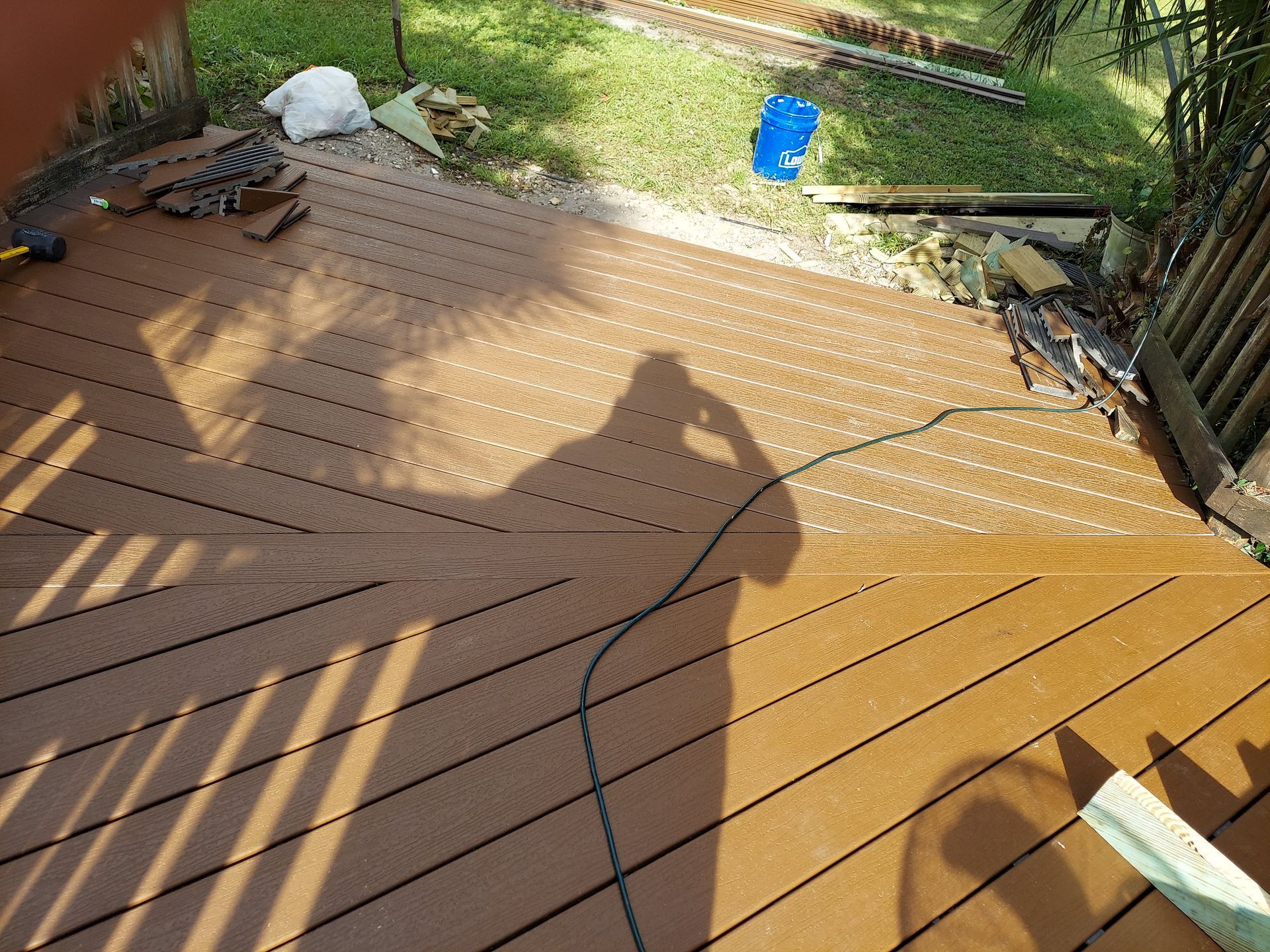 Scuffed wooden deck with cracks, black cables, scattered tools, and a blue bucket in the yard beyond