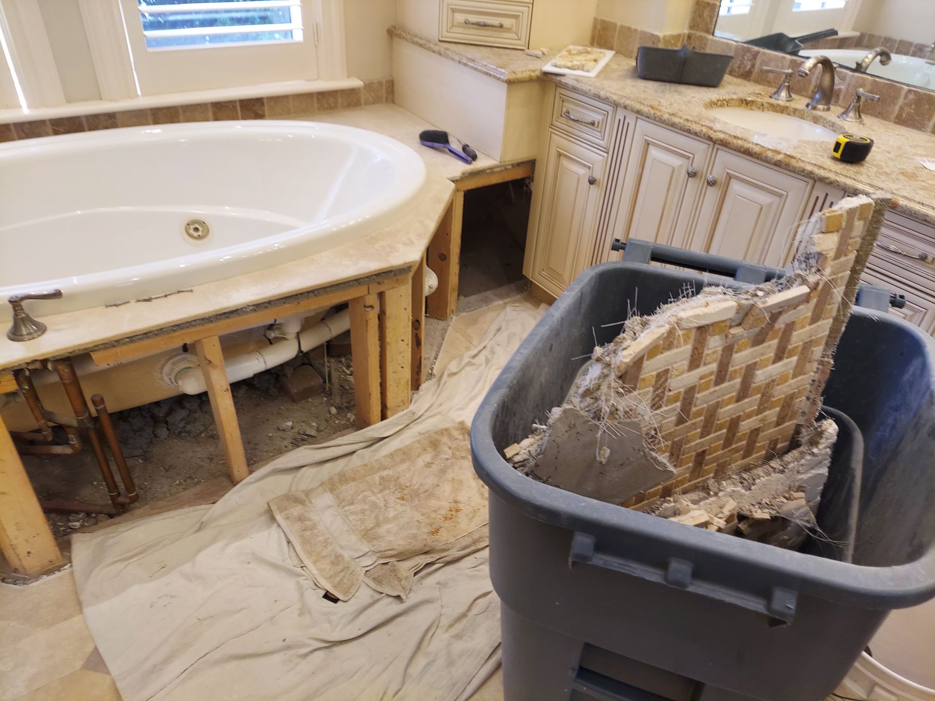 Bathroom under renovation with a built-in tub, exposed framing, and a trash bin full of debris