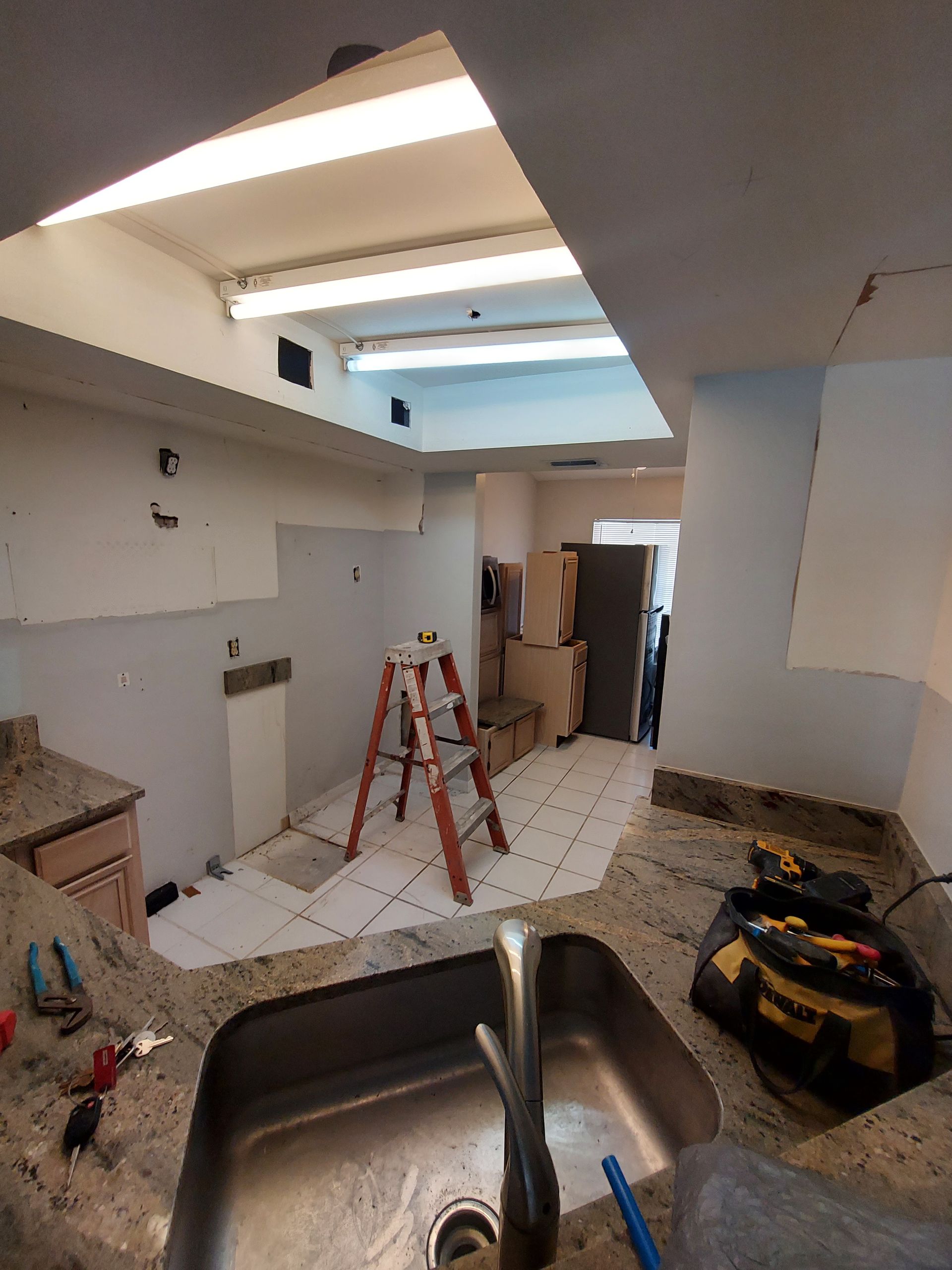 Kitchen under renovation with red step ladder, sink in foreground, debris on floor, and exposed ceiling lights.