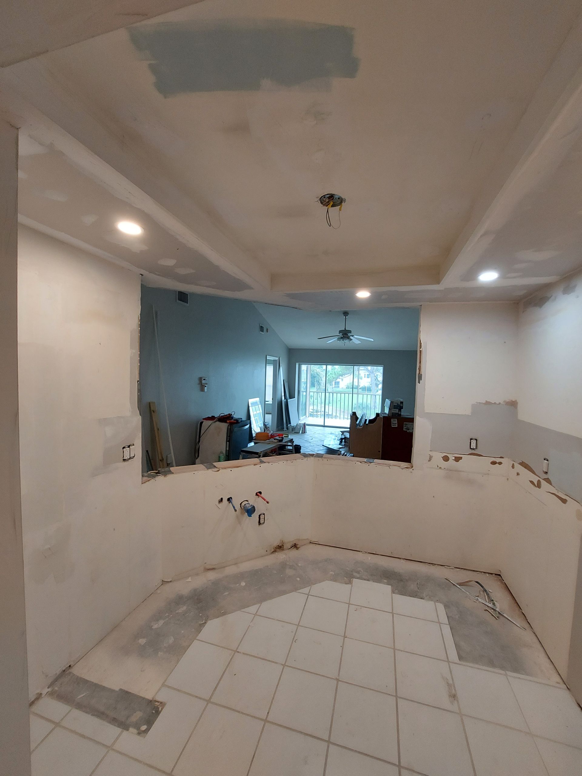 Partially renovated kitchen with white tile floor, blue walls, and unfinished counters leading to a living area