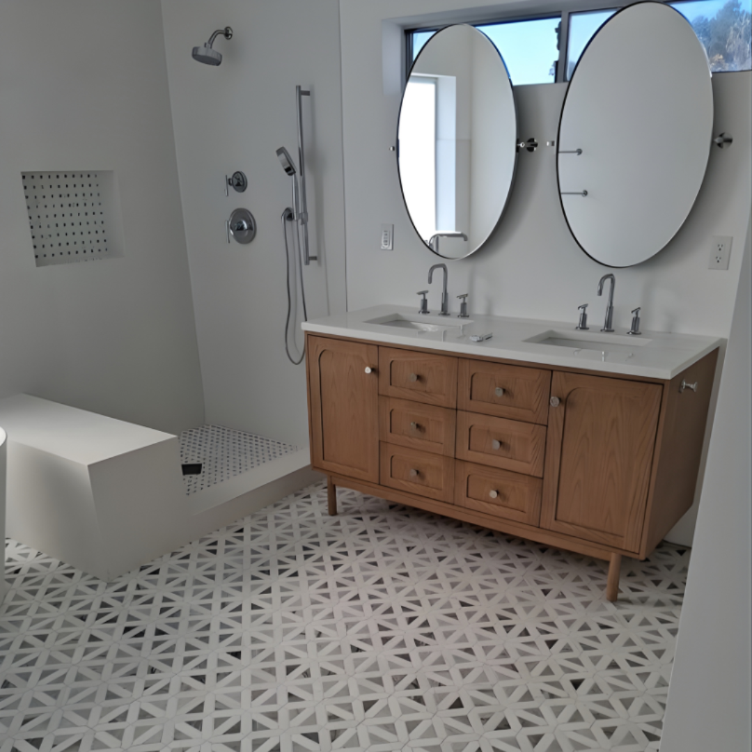 A modern bathroom with a wooden double vanity, two oval mirrors, and a glass-enclosed shower with geometric tile flooring.