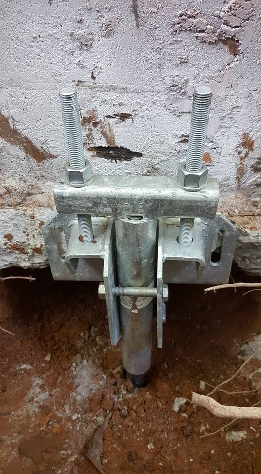 Metal bracket secured to a crumbling brick wall, with a vertical post sunk into the surrounding dirt.